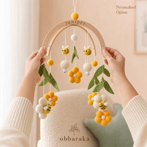 May include: A hanging mobile featuring a curved wooden arch with the name juniper engraved on it. Suspended from the arch are felted wool elements including yellow and white flowers, green leaves, and small bees with black and yellow stripes.