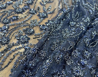 May include: Close-up of navy blue fabric with intricate floral and leaf patterns. The fabric is embellished with sequins and beads in shades of silver and blue, creating a textured, shimmering effect. The design features detailed embroidery, ideal for evening wear or special occasion garments.