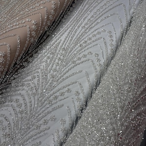 Puede incluir: Primer plano de tres rollos de tela adornada. Las telas son en tonos blanco y beige, con intrincados patrones de cuentas y lentejuelas plateadas. Los diseños incluyen motivos florales y geométricos, creando una apariencia lujosa y texturizada.