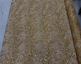 May include: A sheer beige mesh fabric featuring an intricate, repeating vertical pattern of gold sequins and embroidery. The design consists of stylized, branching leaf-like motifs that run the length of the material, creating a dense, shimmering texture.