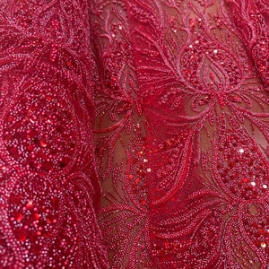 Puede incluir: Primer plano de una tela roja vibrante con intrincados bordados florales. La tela está adornada con lentejuelas y cuentas, creando una apariencia texturizada y lujosa. El diseño presenta patrones detallados y un color rico y saturado.