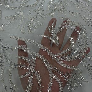 以下が含まれることがあります： 銀色のビーズとスパンコールで装飾された、透明感のある白い生地のクローズアップ。複雑なデザインは、流れるような有機的なパターンを特徴としています。生地は持ち上げられており、光が通り抜け、繊細なディテールと質感を際立たせています。