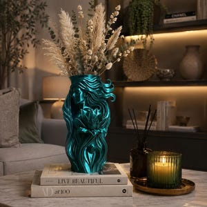 May include: A metallic teal vase featuring a sculpted relief of a woman with flowing hair and a large lotus flower at the centre. The vase is filled with dried pampas grass and eucalyptus stems, resting on two stacked books titled Live Beautiful.