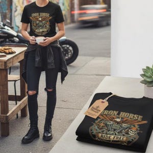 May include: Black t-shirt with a motorcycle graphic and the text "IRON HORSE RIDE FREE". The shirt is folded on a table next to a person wearing the same shirt. The graphic includes an eagle, skull, and motorcycle parts.
