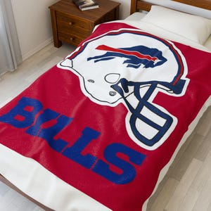 May include: A red and white blanket featuring the Buffalo Bills logo. The blanket displays a white football helmet with blue and red accents, and the word "BILLS" in blue block letters. The blanket is on a bed with a white pillow.