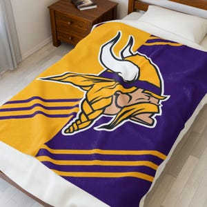 Puede incluir: Una manta de felpa con el logotipo de los Minnesota Vikings. El diseño está dividido diagonalmente, con una sección amarilla que muestra el logotipo de la cabeza vikinga y una sección púrpura con rayas amarillas. La manta está sobre una cama con una almohada blanca.