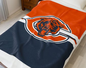 May include: A bedspread with the Chicago Bears logo. The top is orange, with a navy blue section at the bottom. The logo, a bear's head, is in the centre, outlined in white. The bedspread is on a bed with a white pillow.