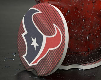 May include: A circular car coaster displaying the Houston Texans logo. The coaster has a red and black dotted pattern with the team's logo in navy blue, red, and white. The coaster is next to a red glass with water droplets.