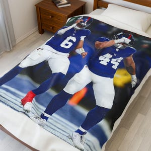 May include: A white blanket featuring two football players in blue uniforms with white pants. The players are in action poses, with one wearing the number 6 and the other wearing the number 44. The blanket is on a bed.