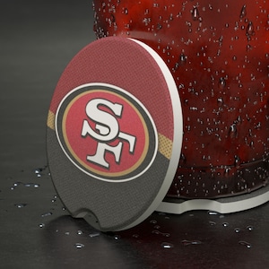 Könnte beinhalten: Ein runder Auto-Untersetzer mit dem Logo der San Francisco 49ers. Der Untersetzer hat ein rotes und schwarzes Design mit dem Logo des Teams in der Mitte. Der Untersetzer befindet sich neben einem Glas mit einem roten Getränk.