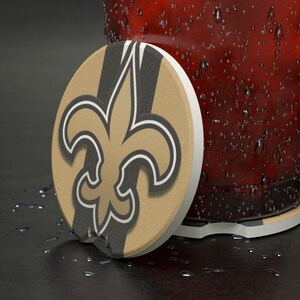 May include: A round drink coaster featuring the New Orleans Saints fleur-de-lis logo in gold, black, and white. The coaster is made of absorbent material and has a white edge. The coaster is next to a glass with condensation.