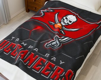 May include: A black blanket featuring the Tampa Bay Buccaneers logo. The logo includes a skull with crossed swords and a football, set against a red background. The team name is displayed in red and white lettering.