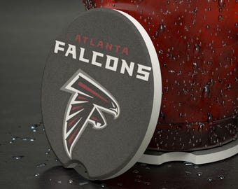 Puede incluir: Un posavasos gris oscuro con el logotipo del equipo de los Atlanta Falcons con el nombre del equipo en letras blancas. El posavasos tiene un borde blanco y un recorte. El logotipo presenta una cabeza de halcón estilizada en rojo y blanco.