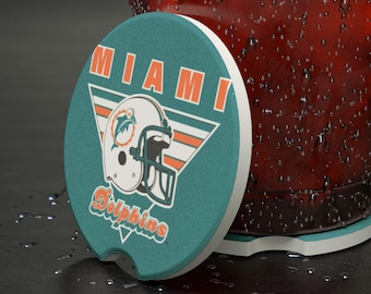 Puede incluir: Un posavasos redondo para coche de color verde azulado y blanco con el logotipo de los Miami Dolphins. El logotipo incluye un casco de fútbol blanco con un delfín naranja y verde azulado, la palabra "MIAMI" en naranja y la palabra "Dolphins" en blanco.
