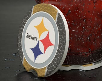 May include: A circular drinks coaster with the Pittsburgh Steelers logo. The coaster has a white border, a gold and grey textured edge, and the Steelers logo in the centre. The logo includes a yellow, red, and blue star. The coaster is next to a glass.