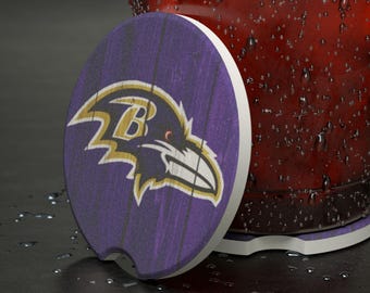 Peut inclure: Un dessous de verre rond pour voiture avec le logo des Ravens de Baltimore. Le dessous de verre a un fond violet imitation bois avec le logo du corbeau de l'équipe en or, noir et blanc. Le dessous de verre est à côté d'un verre avec de la condensation.