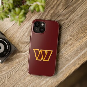 May include: A burgundy phone case with a gold and burgundy "W" logo. The case is on a wooden surface, with a camera and greenery in the background. The phone case is designed for a smartphone.