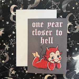 Peut inclure: Une carte gris foncé avec le texte "one year closer to hell" en police gothique blanche et des accents rouges. La carte présente un diable de dessin animé en rouge, allongé. Une carte blanche vierge est derrière.