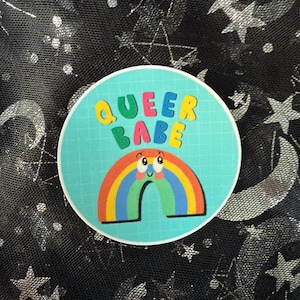 May include: A circular sticker featuring the text "QUEER BABE" in bright, bubble letters above a smiling rainbow. The sticker has a light blue grid background and a white border.
