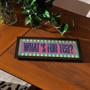 May include: A rectangular framed sign with the words "WHAT'S FOR TEA?" in blue, set against a pink and green background. The sign is in a black frame and rests on a wooden surface, with green plants in the background.