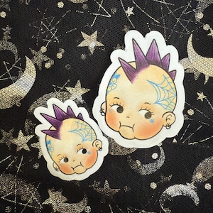 May include: Two stickers featuring a baby with a purple mohawk, blue spiderweb tattoo, and a small earring. The baby has rosy cheeks and a slight frown. The stickers are on a black background with silver stars and moons.
