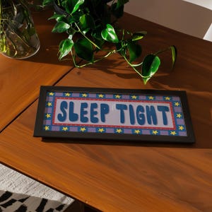 Könnte beinhalten: Ein gerahmtes rechteckiges Schild mit den Worten "SLEEP TIGHT" in blauen Buchstaben. Das Schild hat einen rot-blauen Rand mit gelben Sternen. Der Rahmen ist schwarz. Das Schild befindet sich auf einer Holzoberfläche.