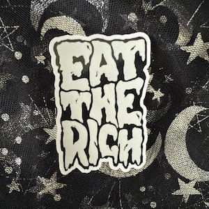 Può includere: Un adesivo bianco con la scritta "EAT THE RICH" in un carattere audace e stilizzato. L'adesivo ha un bordo nero. Lo sfondo è un tessuto nero con un motivo di lune e stelle argentate.
