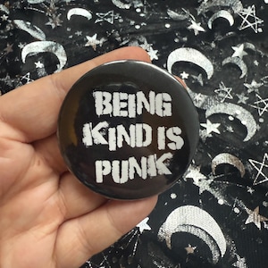 May include: A black button with the words "BEING KIND IS PUNK" in white, distressed lettering. The button is held in a hand, with a black fabric background featuring silver moons and stars.