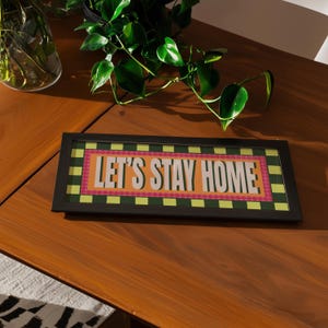May include: A black framed sign with the words "LET'S STAY HOME" in white. The text is on a pink and orange background with a green and yellow checkered border. The sign sits on a wooden table. Green plants are in the background.