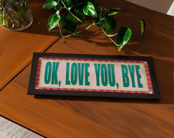 May include: A rectangular framed sign with the text "OK, LOVE YOU, BYE" in green letters. The background is pink with a red and white plaid border. The frame is black. The sign is on a wooden table with green plants in the background.