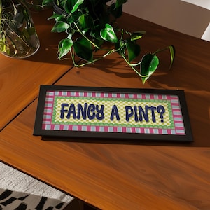 May include: A rectangular framed sign with the text "FANCY A PINT?" in navy blue letters. The sign has a colorful background with a pink, green, and blue checkered pattern. The frame is black. The sign is on a wooden table.