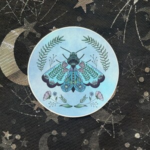 May include: A round sticker with a blue background featuring a detailed illustration of a moth. The moth has intricate wing patterns in shades of blue, green, and pink, surrounded by floral and leaf accents.
