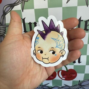 May include: A sticker featuring a baby with a punk rock hairstyle and tattoos. The baby has a purple mohawk, a blue spiderweb tattoo, and a floral tattoo. The sticker is held in a hand against a background with cherries and barbed wire.