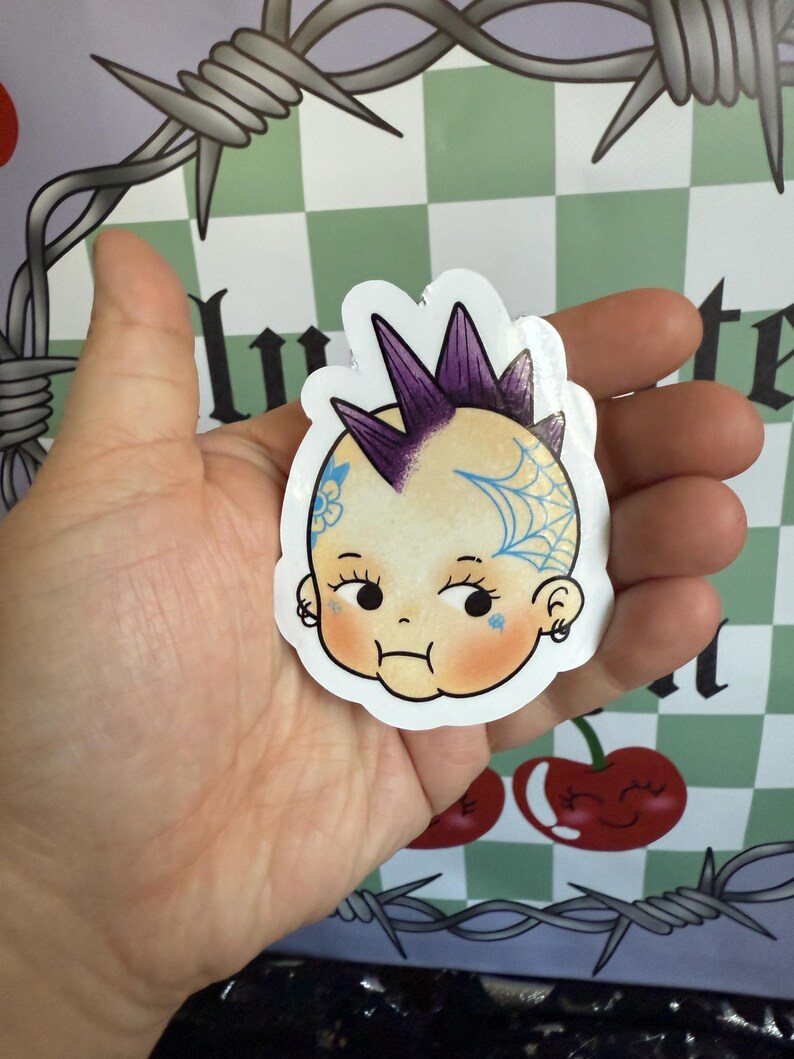 May include: A sticker featuring a baby with a punk rock hairstyle and tattoos. The baby has a purple mohawk, a blue spiderweb tattoo, and a floral tattoo. The sticker is held in a hand against a background with cherries and barbed wire.