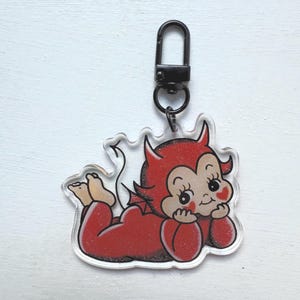 May include: A clear acrylic keychain featuring a cartoon devil in a red outfit with horns, a tail, and a black clasp. The devil is lying down, resting its chin on its hands, with a smiling expression.