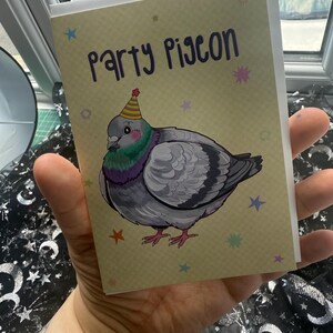 May include: Greeting card featuring a cartoon pigeon wearing a party hat. The text "Party Pigeon" is written in purple script above the illustration. The background is tan with colorful star accents. The card is held in a hand.