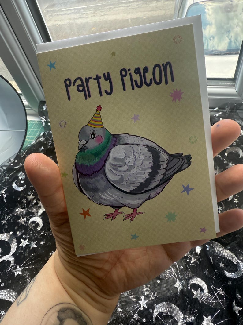 May include: Greeting card featuring a cartoon pigeon wearing a party hat. The text "Party Pigeon" is written in purple script above the illustration. The background is tan with colorful star accents. The card is held in a hand.