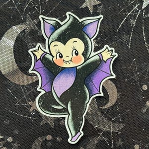 Op de afbeelding: Een grillige sticker met een cartoon vleermuis karakter in een speelse pose. De vleermuis is zwart met paarse vleugels en accenten, en heeft een schattige, lachende uitdrukking. De achtergrond is een donkere, glinsterende stof met zilveren manen en sterren.