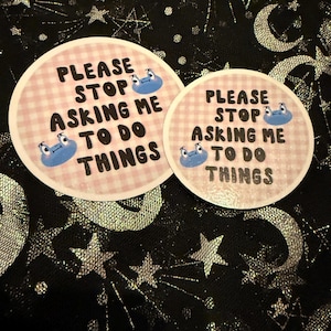 May include: Two round stickers with the text "PLEASE STOP ASKING ME TO DO THINGS" in black font. The stickers have a pink gingham pattern background and blue cartoon frog illustrations.