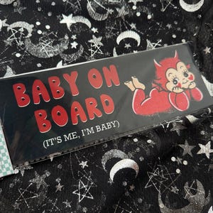 May include: A rectangular black sign with red text reading BABY ON BOARD and (IT'S ME, I'M BABY) in white. A red cartoon devil character with horns and a tail is positioned on the right side, resting on its stomach with its chin in its hands.
