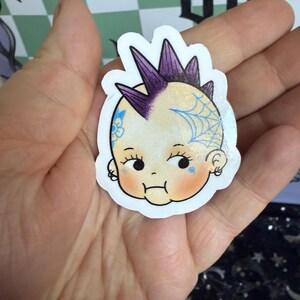 May include: A sticker featuring a cartoon baby with a purple mohawk and a blue spiderweb tattoo. The baby has rosy cheeks and a small frown. The sticker is held in a hand.