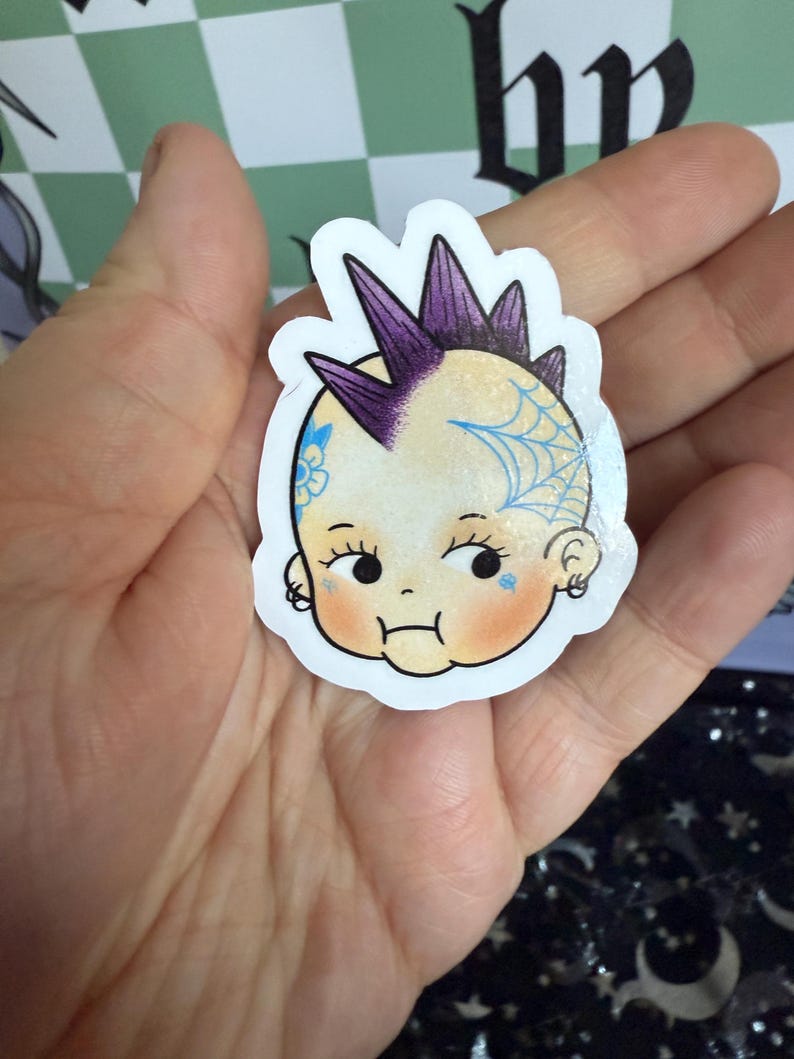 May include: A sticker featuring a cartoon baby with a purple mohawk and a blue spiderweb tattoo. The baby has rosy cheeks and a small frown. The sticker is held in a hand.