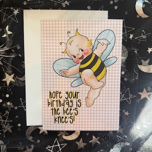 May include: A greeting card with a cartoon bee illustration. The bee is wearing a black and yellow striped outfit with blue wings. The card has the text "hope your birthday is the bee's knees!" on a pink and white checkered background.