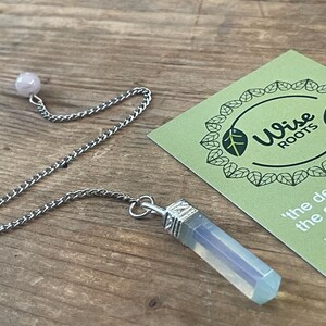 Puede incluir: Un péndulo de cristal con una cadena plateada y un cristal hexagonal azul claro. Una pequeña esfera rosa está en la parte superior de la cadena. Una tarjeta verde con el texto "Wise Roots" está en el fondo.