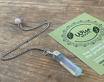 Op de afbeelding: Een kristallen slinger met een zilveren ketting en een lichtblauwe zeshoekige kristal. Bovenop de ketting zit een kleine roze bol. Een groene kaart met de tekst "Wise Roots" staat op de achtergrond.