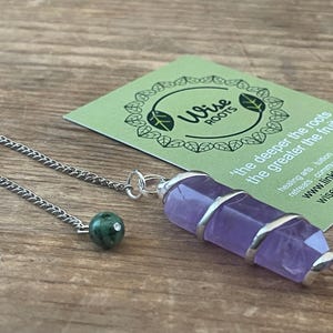 Puede incluir: Un péndulo de cristal de amatista púrpura con una cadena plateada y una cuenta verde. El péndulo está junto a una tarjeta verde con el texto "Wise Roots" y "the deeper the roots the greater the fruits".