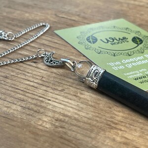 Può includere: Un pendolo d'argento con un cristallo nero e una sfera di cristallo trasparente. La catena ha un ciondolo a forma di sole e un ciondolo a forma di luna crescente. Sullo sfondo una carta verde con la scritta "Wise Roots".