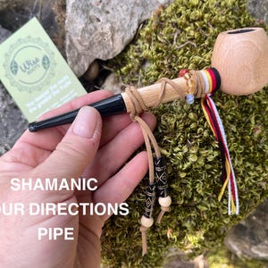 Puede incluir: Una pipa de madera con un hornillo de color claro y una boquilla negra. La boquilla está envuelta en un cordón de cuero beige con dos cuentas colgantes con patrones en blanco y negro. Una cinta roja, amarilla, negra y blanca está unida al hornillo.