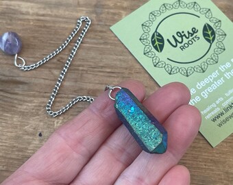 Op de afbeelding: Een pendel met een zilveren ketting en een blauwgroene kristallen punt. Een kleine, ronde, paarse steen is aan de ketting bevestigd. Een groene kaart met de tekst "Wise ROOTS" staat op de achtergrond.