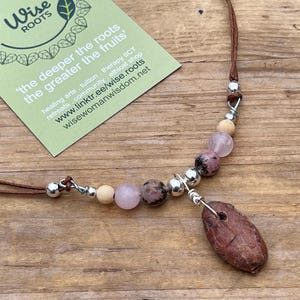 May include: A handcrafted necklace with a brown leather cord, silver beads, and pink, beige, and brown stone beads. The necklace has a pendant and a tag with the text "Wise Roots".